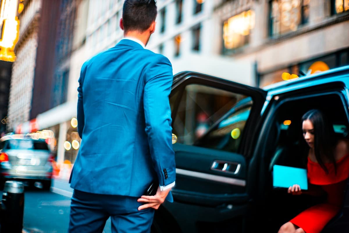A chauffeur in a suit opens the rear door of a sedan for his passenger